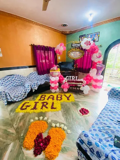 A living room setup for a 'Welcome Baby Girl' event, with flower rangoli, balloon pillars, and a decorated cradle area.