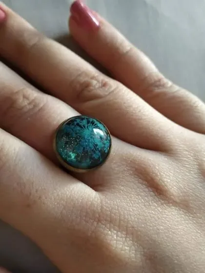 This ring showcases a beautiful blue-green flower with a sparkling, galaxy-like effect. The dark background makes the colors appear deep and vibrant.