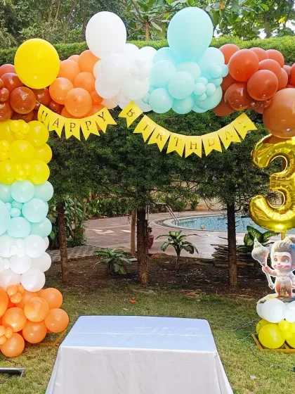 An outdoor birthday setup with a Hanuman theme. The colorful balloon arch in orange, yellow, and blue is perfect for a poolside party, complete with a small Hanuman cutout.