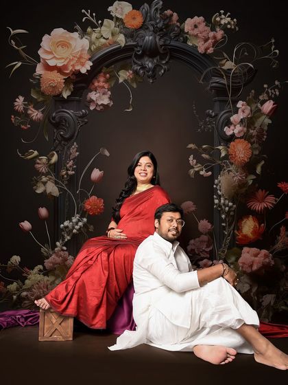 A regal portrait of an expecting couple against a dark, ornate floral backdrop. The mom-to-be's red saree and her partner's white kurta create a striking and elegant contrast.