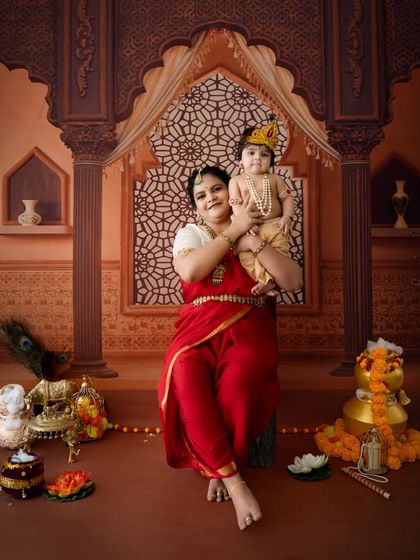 A regal portrait of a mother holding her little Krishna, who is wearing a crown, in front of an ornate archway backdrop.