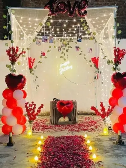 An outdoor romantic setup for a birthday or anniversary. It features a canopy with fairy lights, a rose petal aisle, and red and white balloon pillars.