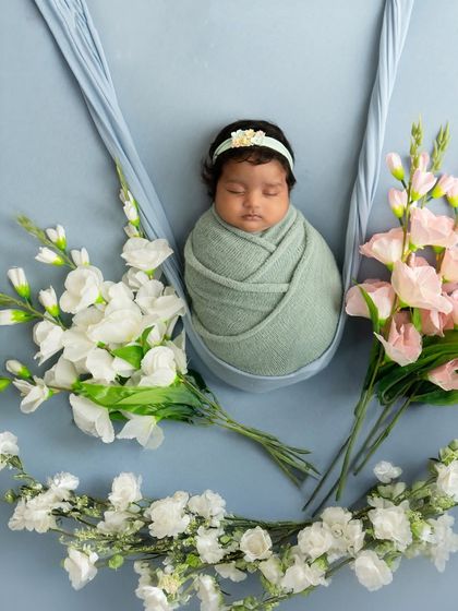 A serene portrait of a baby swaddled in mint green, resting in a fabric swing and framed by bouquets of white and pink flowers.