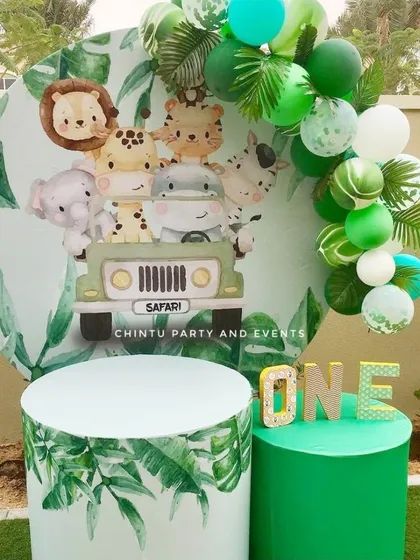 An outdoor jungle safari themed first birthday. The setup includes a round backdrop with cute animals, themed pedestals, and a green balloon garland.