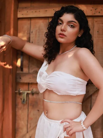 A confident and direct pose from the Apsara series. The lighting highlights the model's features and the texture of the white bustier top and ruffled skirt.
