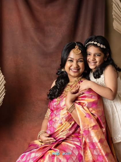 An elegant maternity portrait celebrating Indian tradition. The mother-to-be wears a beautiful pink saree, joined by her daughter in a lovely studio setup with boho elements.