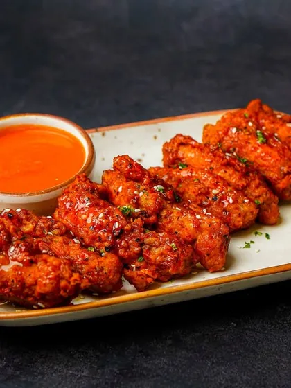 A platter of spicy chicken wings coated in a glossy red sauce, ready to be dipped. The dark, dramatic lighting highlights the texture of the wings, making them look extra crispy and saucy.