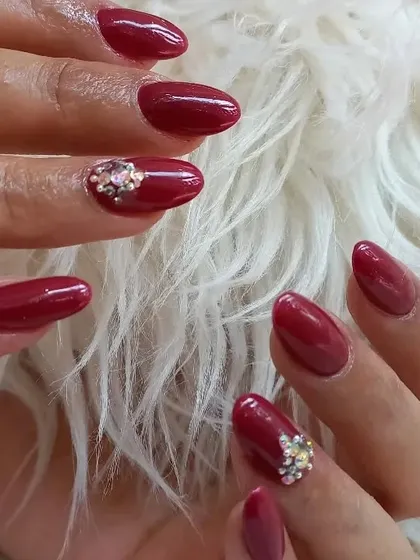 A classic deep red manicure on oval nails, elevated with a beautiful cluster of iridescent rhinestones at the base of the ring finger nail for a touch of elegant sparkle.