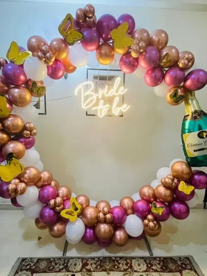 A whimsical "Bride to be" balloon ring in shades of rose gold, magenta, and white. The decoration is accented with butterfly cutouts and a champagne bottle foil balloon, creating a playful and feminine party vibe.