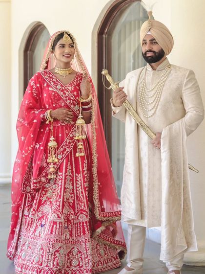 A regal portrait of a Sikh couple on their wedding day. The groom holds the ceremonial sword (kirpan), and the bride showcases her traditional red lehenga and kalire.