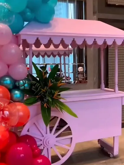 A charming pink cart prop, perfect for a dessert station or candy bar at a children's party. It's decorated with a vibrant balloon garland.