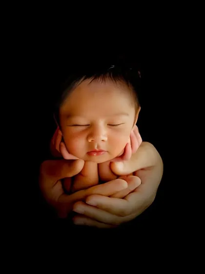 A dramatic low-key portrait focusing on the baby's face cradled in hands. This artistic shot emphasizes the emotion and connection in a powerful way.