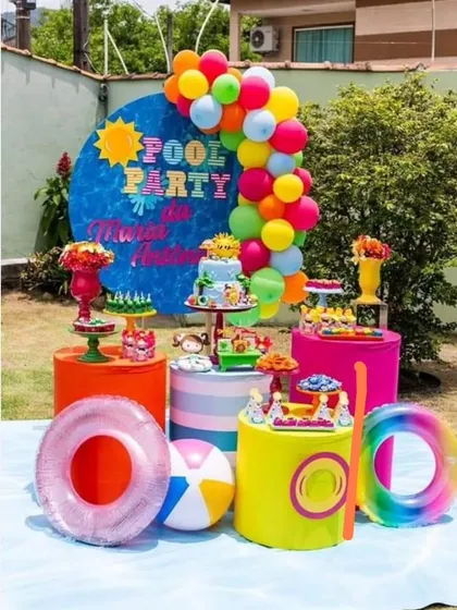 A vibrant and fun poolside birthday party. The decor is full of bright colors, with a large "Pool Party" backdrop, balloon arch, and themed props.
