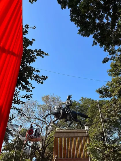 A low-angle shot of the Shivaji Maharaj statue against a clear blue sky, framed by a red flag. This composition creates a powerful and patriotic image.