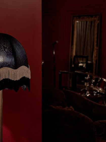 A custom-designed lamp with a fringed, black patterned shade stands against the rich red wall. This diptych highlights the moody lighting and intimate details that contribute to the bar's sophisticated, old-fashioned vibe.