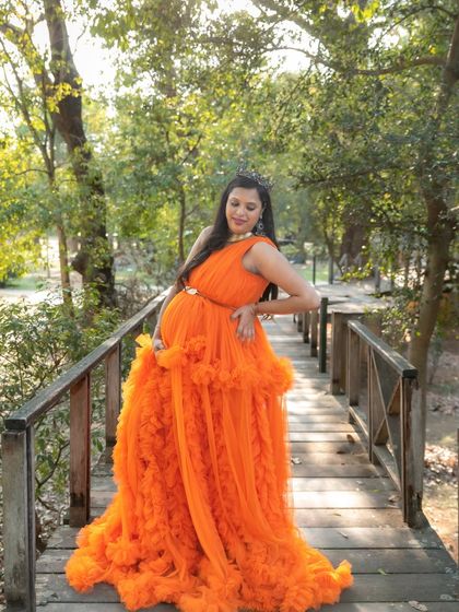 A regal portrait of a mom-to-be in an orange gown and tiara, posing on a wooden bridge in a park.