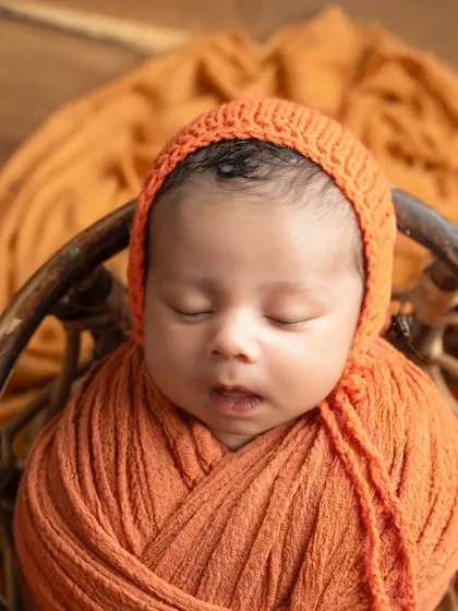 A close-up of the sleeping newborn's face, showing the texture of the knitted orange bonnet and their peaceful expression.