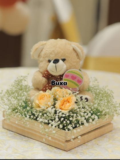 A classic teddy bear centerpiece with a custom rattle cutout, surrounded by delicate flowers in a wooden crate.