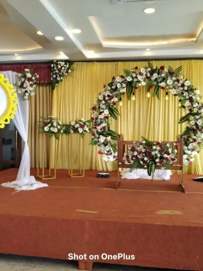 A naming ceremony setup with a red carpeted stage. The cradle is framed by a circular arch of red and white flowers, with a decorative mirror element adding a unique touch to the side.
