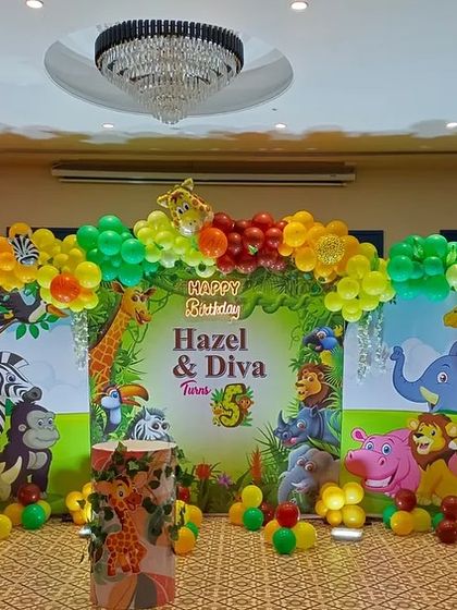 A lively jungle-themed 5th birthday party for twins. The stage backdrop is filled with cartoon animals, and a colorful balloon arch completes the look.