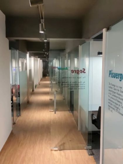 A view down the main corridor of the Amigo co-working office. Glass-walled private cabins line the hallway, each named after a river. This maintains a sense of openness and community while providing distinct, private workspaces.