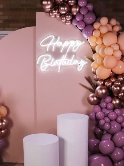 Modern and chic. This birthday setup uses a pink arched backdrop, a "Happy Birthday" neon sign, and an artistic balloon arrangement in purple, peach, and rose gold for a trendy, stylish look.