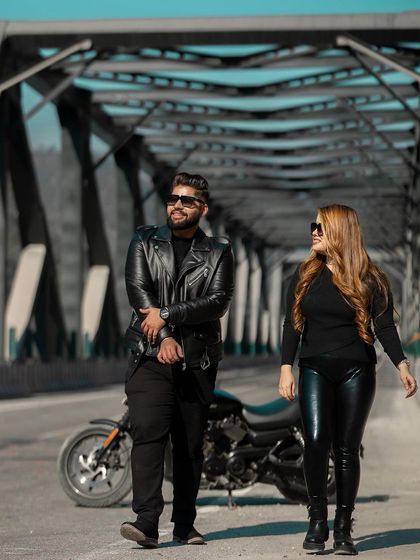 A stylish shot of the couple walking on a bridge with their motorcycle. The coordinated all-black outfits and confident poses create a modern and fashionable pre-wedding photograph.