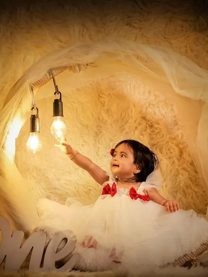 A magical first birthday photo, where the little girl reaches for glowing lightbulbs in a dreamy, warm-toned setup.