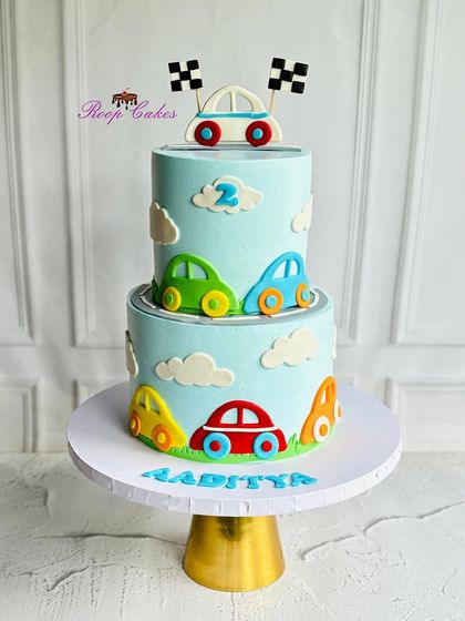 A fun two-tier cake for a little car enthusiast turning two. The light blue cake is decorated with a winding road and colorful fondant cars, topped with a race car and checkered flags.