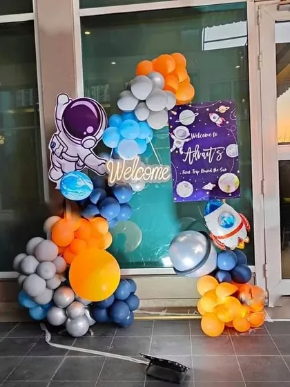 A welcome setup for a space-themed party. This arrangement includes an astronaut cutout, a personalized welcome sign, and a balloon garland in orange, blue, and grey.