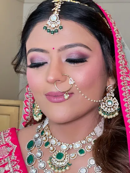 A close-up of a stunning bridal makeup look. The focus is on the intricate smokey eye with shades of pink and purple, paired with a matte pink lip and glowing skin, perfectly complementing her green and pink jewelry.