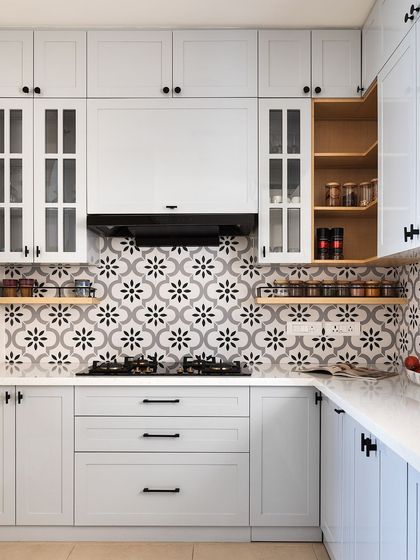 This kitchen has a classic, timeless feel with its white shaker-style cabinets and black hardware. The monochrome floral-patterned backsplash adds a touch of personality, and open wooden shelves provide easy access to spices.