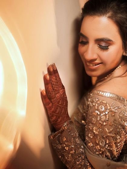 A soft, artistic portrait of the bride. A light flare adds a dreamy quality to the image as she smiles, her hand with intricate henna resting gently against the wall.