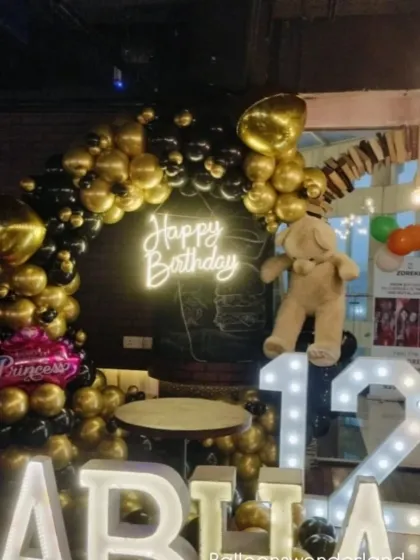 A stylish black and gold theme for a 12th birthday. This setup includes a balloon arch, a 'Happy Birthday' neon sign, light-up marquee letters and numbers, and a cute teddy bear accent.