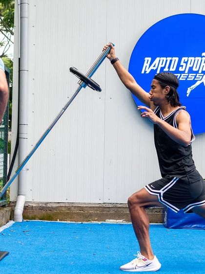 An athlete performs a landmine press, a versatile exercise for developing upper body pushing strength and shoulder stability in a joint-friendly manner. The coach's watchful eye ensures proper form.