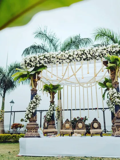 An elegant outdoor mandap blending traditional South Indian elements with a clean, modern aesthetic. The white floral canopy and carved pillars stand beautifully against the lush greenery and palm trees.
