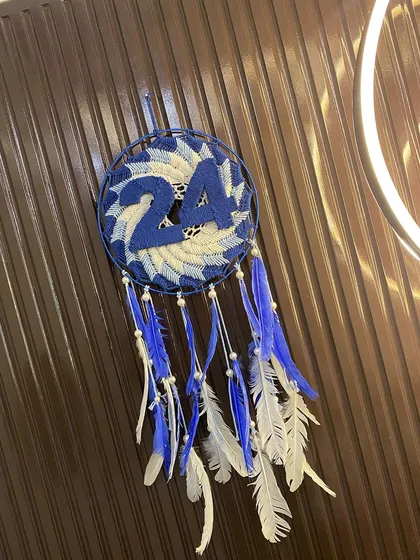 The finished number 24 numerology dreamcatcher, hung against a dark background. The blue and white design with hanging feathers is believed to bring luck and prosperity.