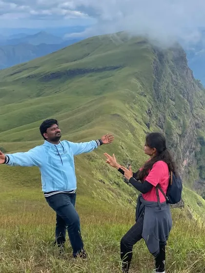 A fun, candid moment between two trekkers with the stunning Nethravathi peak in the background.