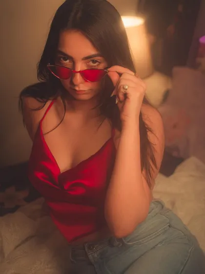 A playful and confident shot in a red satin top, peering over red sunglasses.