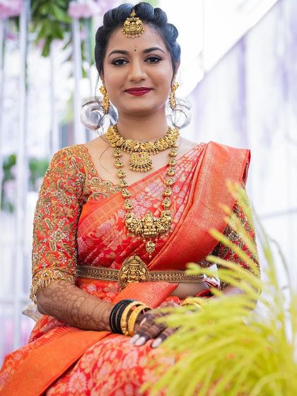 A stunning bridal portrait in a vibrant orange silk saree. This look features classic winged eyeliner and a bold red lip, paired with heavy temple jewellery for a truly traditional South Indian wedding style.