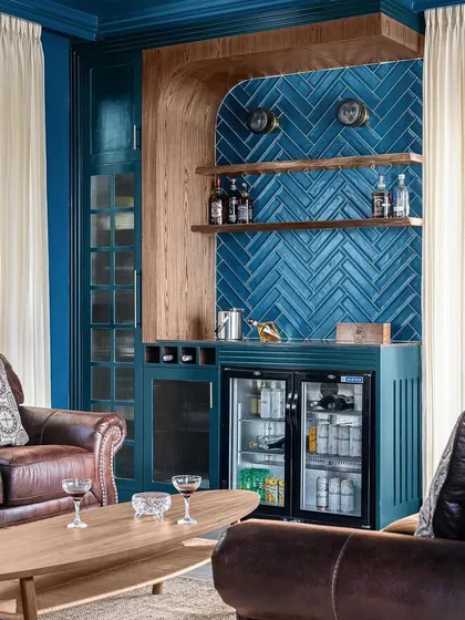 A full view of the teal entertainment room. The bar unit is nestled in a custom arched nook with a herringbone tile backsplash. The rich color and warm wood create a sophisticated space for hosting.