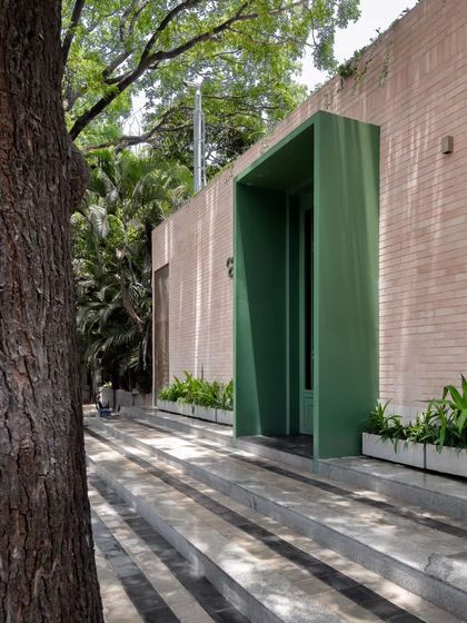 An angled view of the restaurant's entrance, highlighting the deep green portal that draws you in from the street. The surrounding greenery and natural stone steps create a gentle transition from the urban environment to the calm interior.