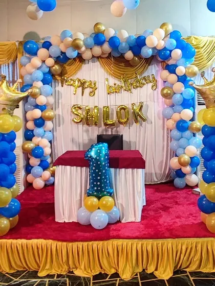 A royal-themed first birthday decoration for a boy. The setup features a blue, gold, and white balloon arch on a small stage, with two large balloon columns topped with gold foil crowns.