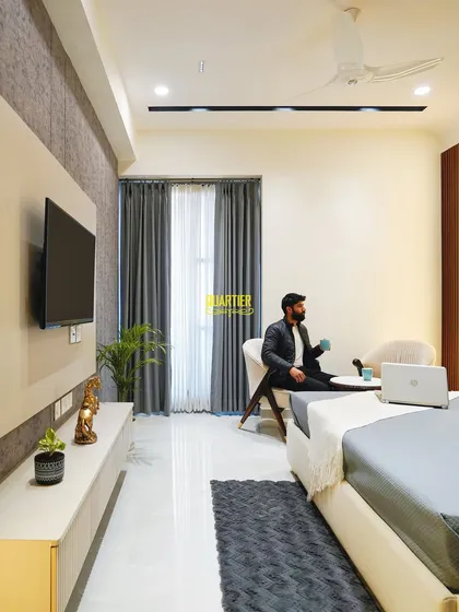 This parents' bedroom at Apex D'Rio is designed as a modern sanctuary. The space includes a comfortable seating area and a sleek, floating TV unit, all set against a backdrop of textured wallpaper and wood-look louvers.