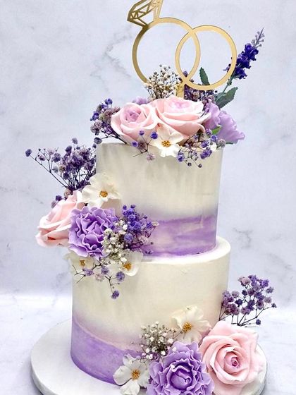 A beautiful two-tiered engagement cake featuring a soft purple watercolor wash. It's adorned with fresh roses, carnations, and baby's breath, and topped with a custom gold ring topper to celebrate the promise of forever.