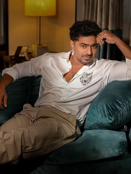 A relaxed portrait of actor Dev Adhikari on a couch. The soft, ambient lighting and his casual pose create a comfortable and approachable feel, contrasting with his more intense looks.