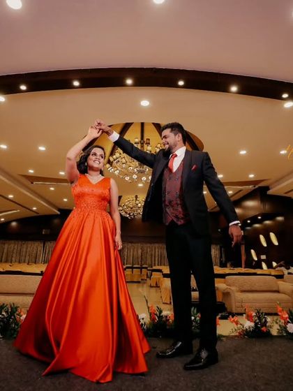 A wide shot of the couple sharing their first dance at the reception. The bride's flowing orange gown and the grand ballroom setting create a cinematic feel.