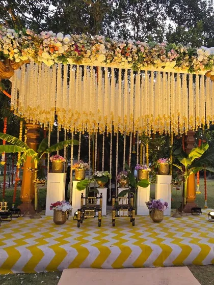 A stunning South Indian theme Haldi stage. The mandap is built with traditional wooden pillars, a canopy of jasmine flowers, and a vibrant yellow and white chevron-patterned floor.