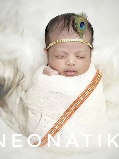 A minimalist and modern take on the newborn Krishna theme, with the baby wrapped in white on a fluffy surface, accented by a single peacock feather.