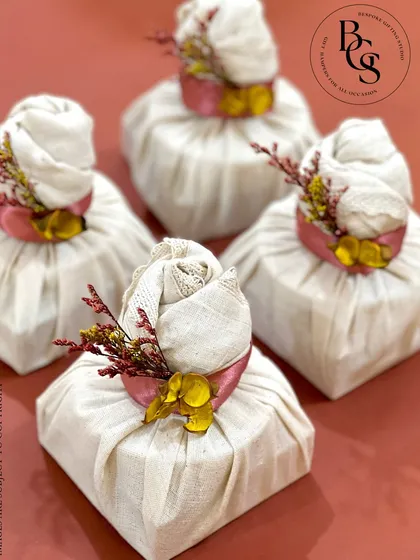 An eco-friendly and rustic wrapping style. These small gifts are wrapped in natural cream-colored fabric using a 'potli' or bundle technique, tied with a pink ribbon and accented with dried flowers.